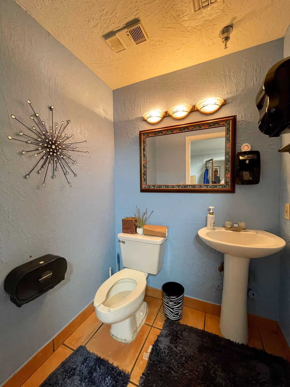 A small bathroom with light blue textured walls and a tiled floor. The room contains a white toilet with the lid up, a white pedestal sink with a soap dispenser, a rectangular mirror with a decorative frame above the sink, and a black trash can with a zebra pattern. There are two black wall-mounted dispensers, one for paper towels and one for soap. A decorative starburst wall art is mounted on the left wall. The ceiling has a vent and a light fixture with three bulbs above the mirror.
