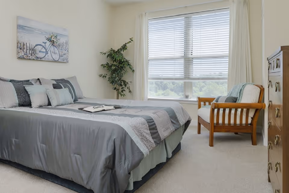 A bright bedroom featuring a made bed with gray bedding, a wooden armchair by a large window, a dresser, and wall art.