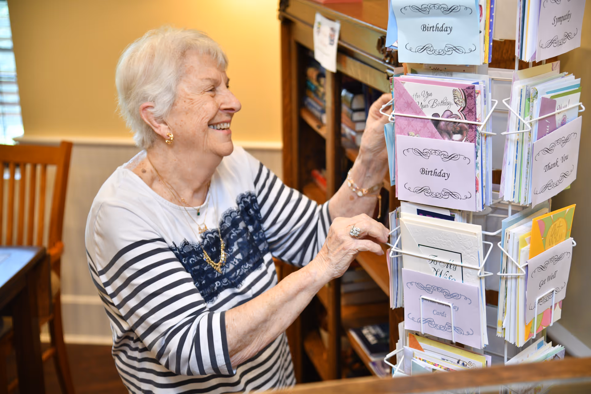 An elderly woman with short white hair and a striped shirt is smiling while selecting a greeting card from a rotating card display rack inside a warmly lit room with wooden furniture.