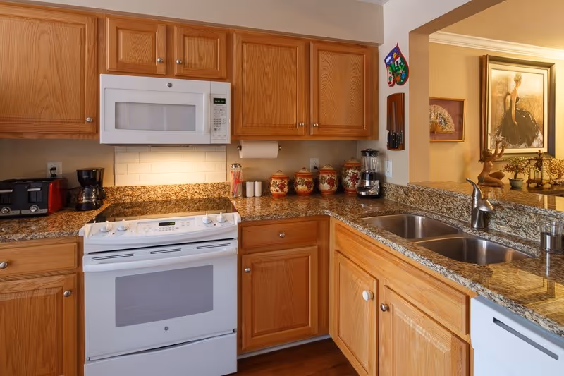 A kitchen with wooden cabinets, granite countertops, a white stove with an oven, a white microwave above the stove, a double sink with a modern faucet, a toaster, a coffee maker, a blender, and decorative canisters on the counter. In the background, there is a partial view of a living area with artwork and decorative items.