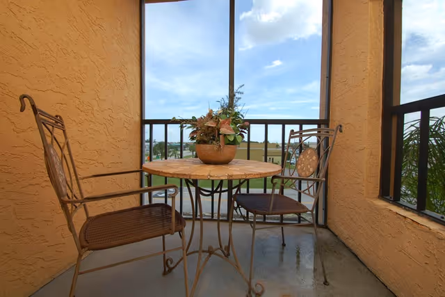 A small outdoor balcony area with a round metal table and two matching metal chairs. A potted plant with flowers is placed on the table. The balcony has a railing and offers a view of a grassy area and buildings in the distance under a partly cloudy sky.