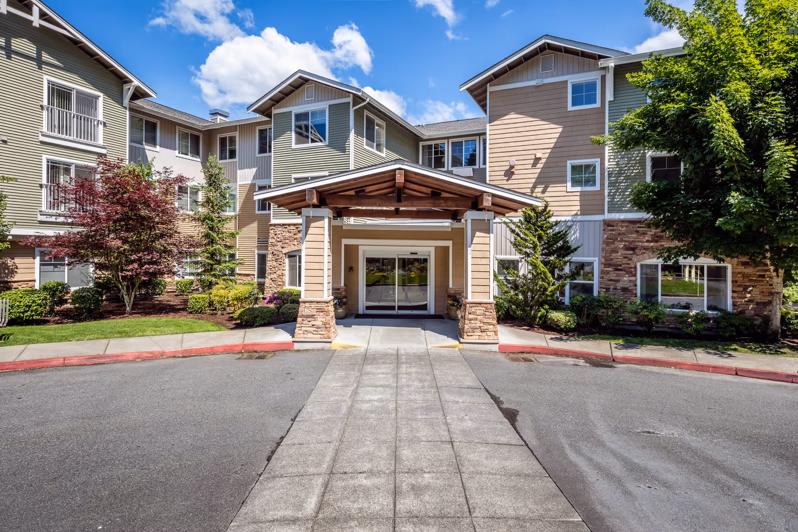 Front exterior view of Woodland Terrace senior living facility showing a three-story building with beige and green siding, stone accents, and a covered entrance. The entrance is flanked by small trees and shrubs, with a clear blue sky and some clouds in the background.