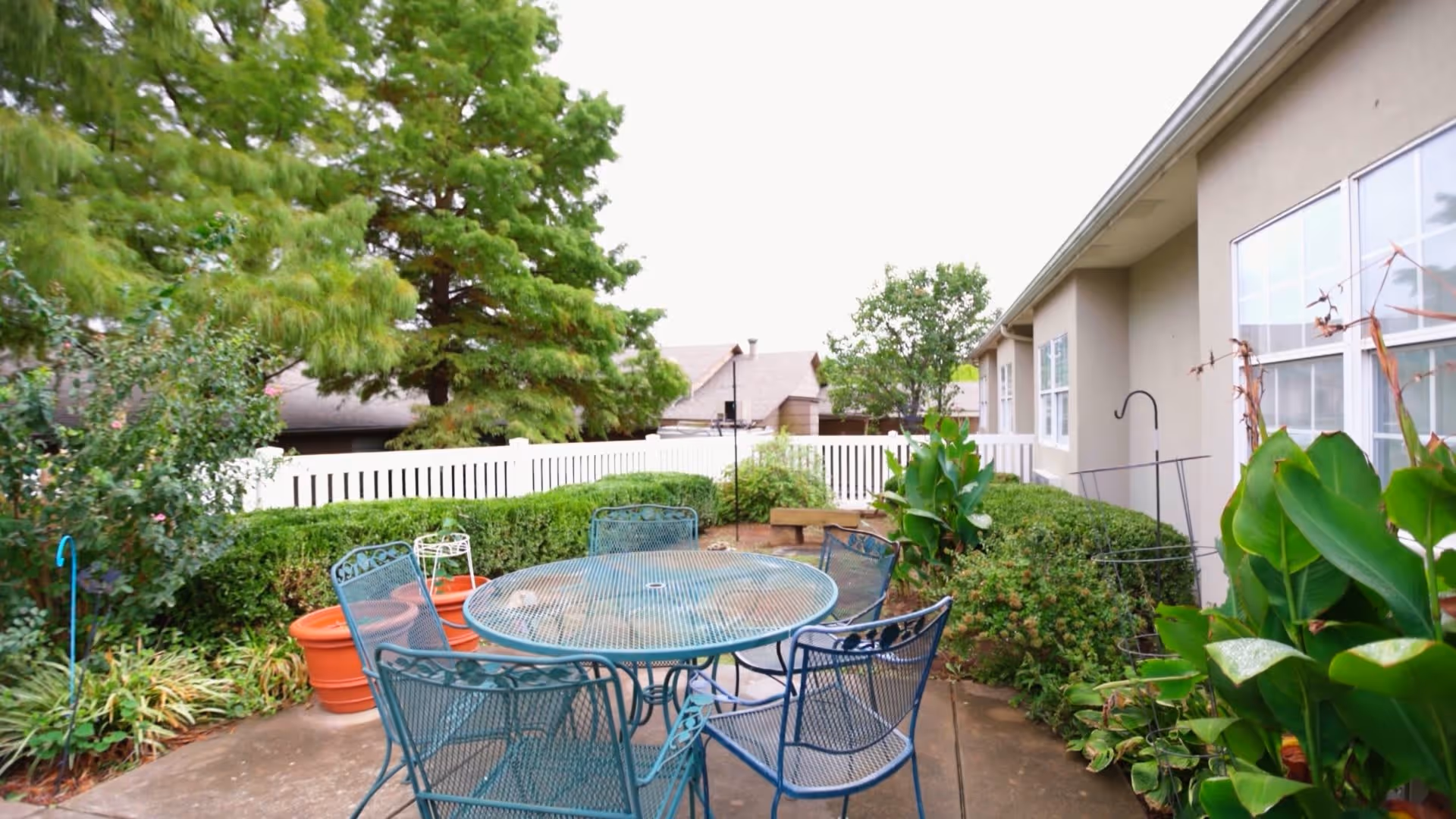 Outdoor patio area with a round metal table and six matching chairs surrounded by green plants and bushes. A white fence and trees are visible in the background next to a beige building with windows.