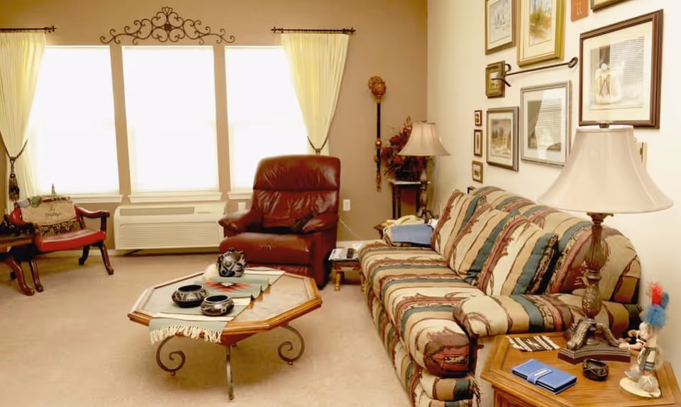 A cozy living room with a patterned sofa, a brown leather recliner, and a wooden coffee table with decorative items. The room has beige walls adorned with framed pictures and two windows with yellow curtains letting in natural light. There are two side tables with lamps and various small decorations.
