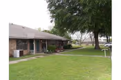 Single-story brick residential building with a covered porch, surrounded by a well-maintained lawn and large trees, with a sidewalk leading to the entrance.