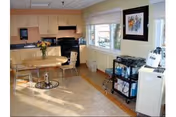 Small communal dining area with a kitchenette, round table and chairs, a window with framed artwork, and a utility cart with equipment.
