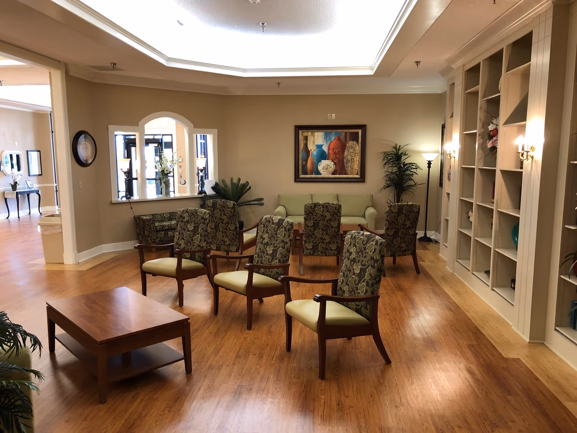 A well-lit living room area with wooden flooring, featuring a set of six patterned armchairs arranged around a wooden coffee table. There is a green sofa against the far wall beneath a framed painting of vases. The room has built-in shelves on the right side with decorative items and plants, and a floor lamp in the corner. An arched window opening looks into another room with additional seating and decor.