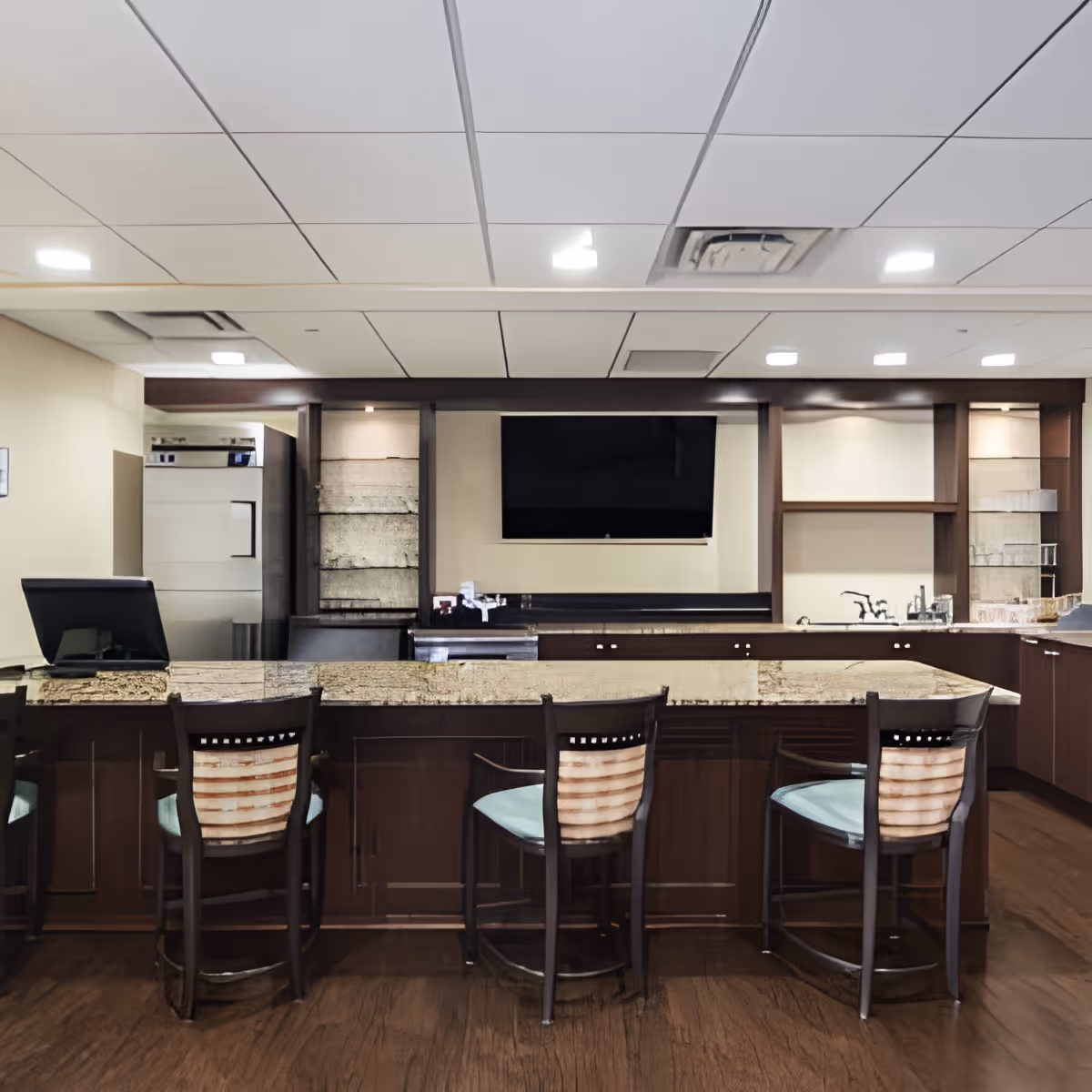 Interior view of a kitchen area in a senior living facility featuring a long granite countertop with four wooden chairs with cushioned seats. The background includes built-in cabinetry, a sink, and a mounted flat-screen TV on the wall. The ceiling has recessed lighting and a drop ceiling design.