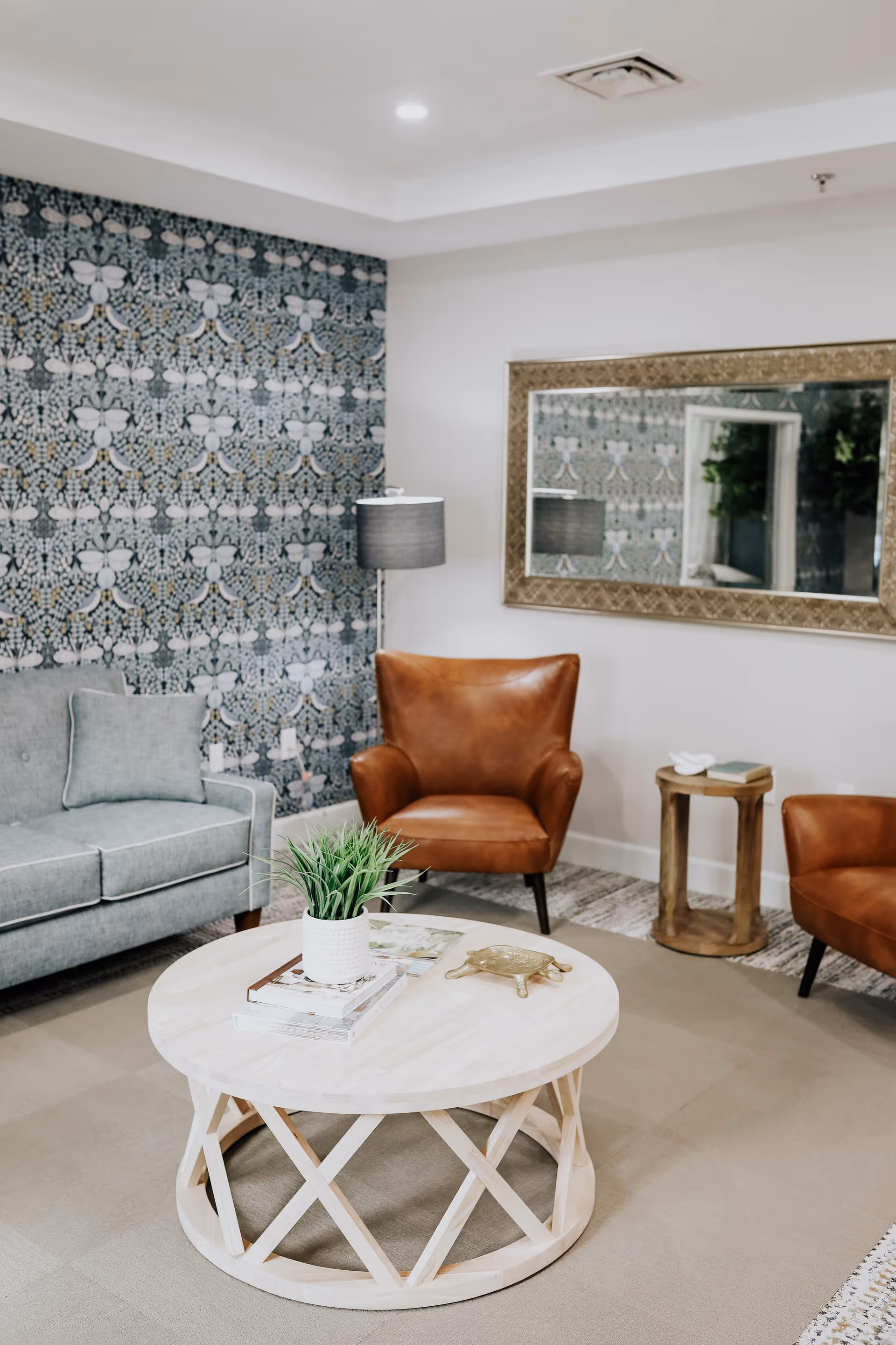 A cozy living room area with a light gray sofa, two brown leather armchairs, a round wooden coffee table with a potted plant and books on it, a floor lamp, and a large decorative mirror on the wall. One wall features a patterned wallpaper with a floral design.
