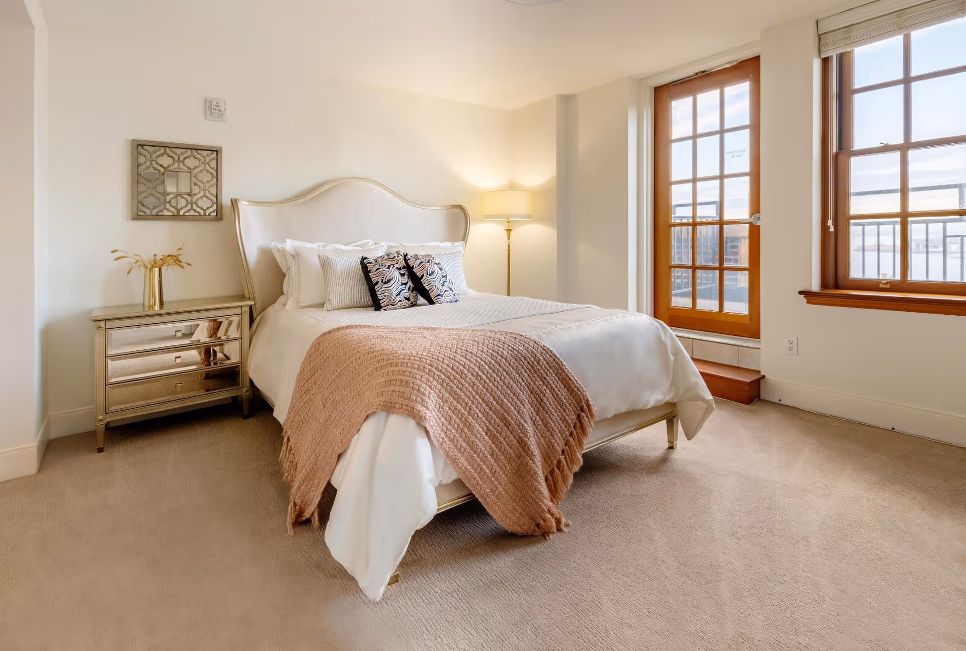 A bright, furnished bedroom with a bed, mirrored nightstand, floor lamp and French doors/windows letting in natural light.