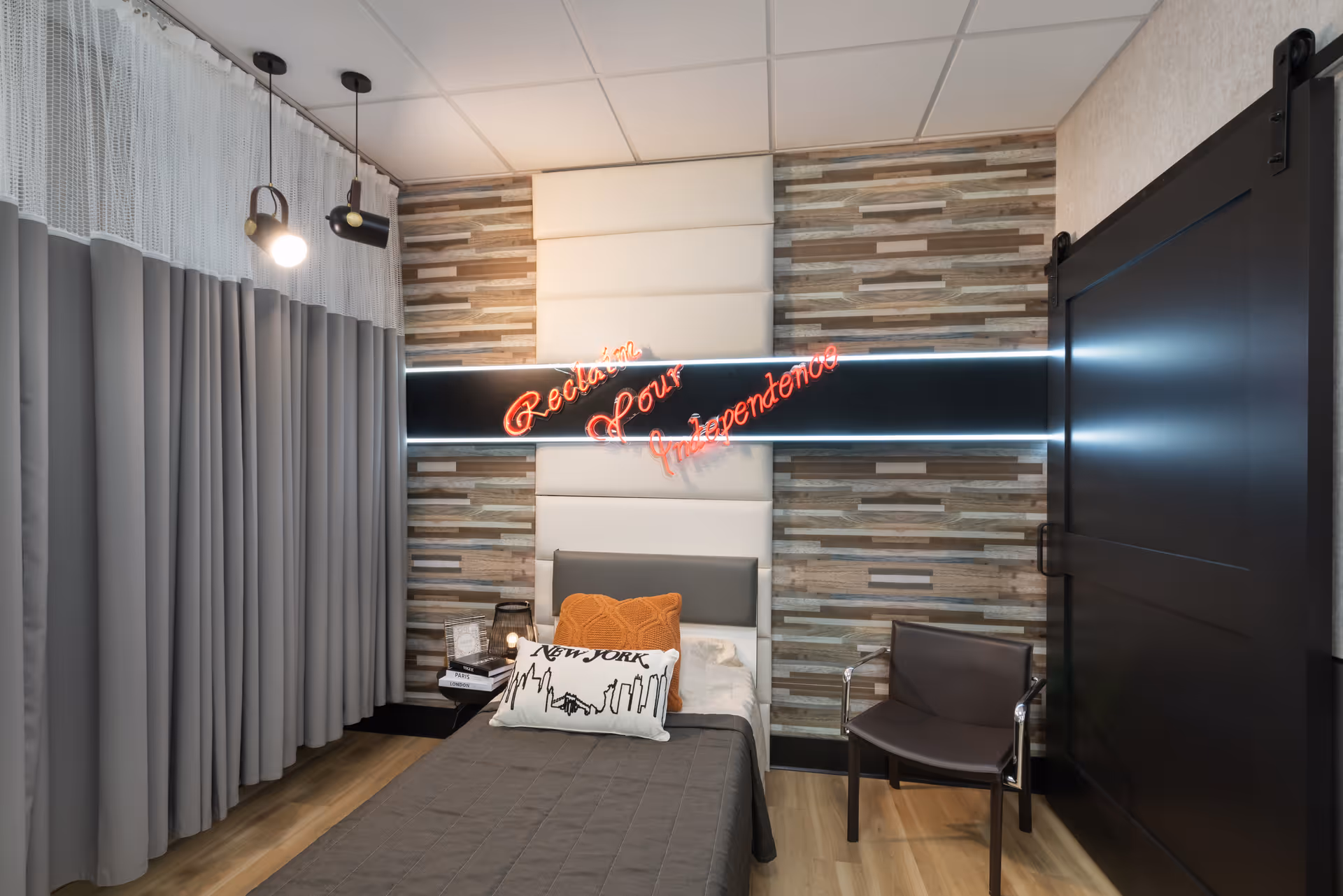 A modern bedroom with a single bed covered in a gray quilt and two decorative pillows, one orange and one white with a New York skyline design. The wall behind the bed features a textured wood panel design with a padded white section and a neon sign that reads 'Reclaim Your Independence'. There are two hanging lights on the left side above gray curtains, a small side table with books and a lantern, and a dark sliding barn door on the right side. A brown chair is positioned next to the bed.