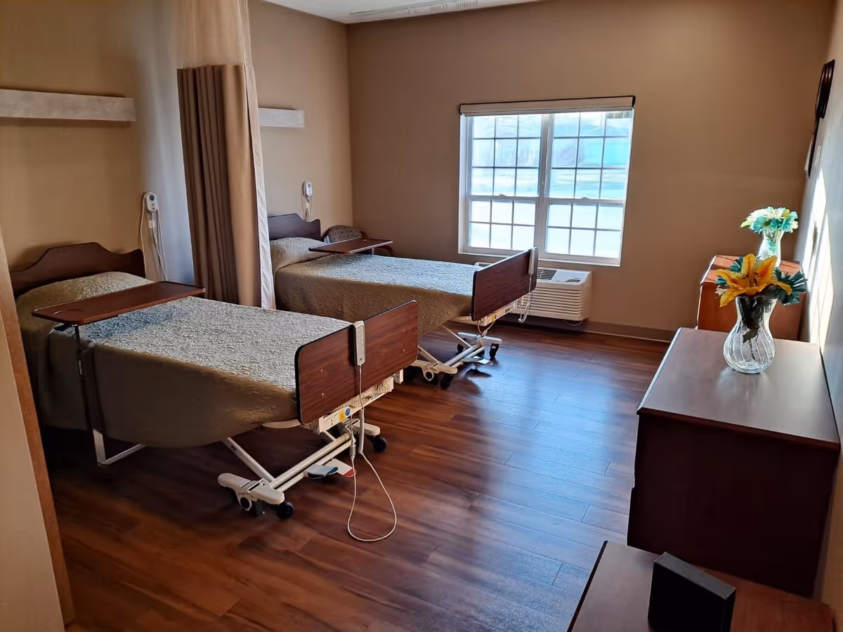Two hospital beds in a shared patient room with curtains, window, and a dresser with flowers.