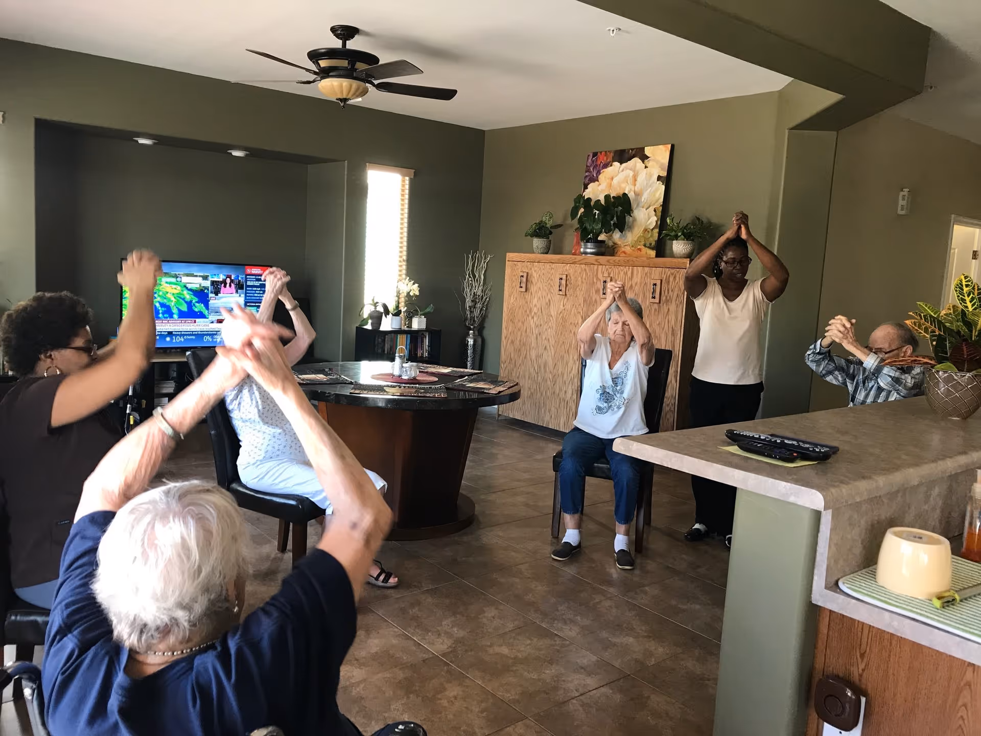 A group of elderly people and a caregiver sitting in a living room area participating in a seated exercise session with their hands raised and clasped above their heads. The room has green walls, a ceiling fan, a television, and a round table with chairs.