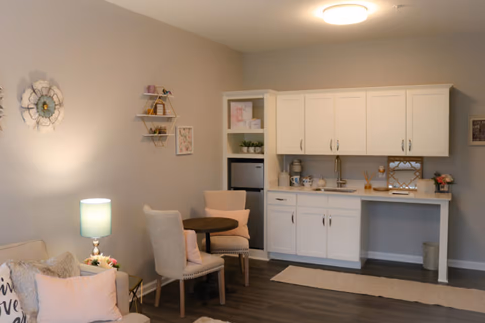A cozy interior space featuring a small kitchenette with white cabinets, a sink, and a mini refrigerator. In front of the kitchenette is a small round table with two upholstered chairs. To the left, there is a beige sofa with decorative pillows and a side table with a lamp. The walls are decorated with floral wall art and shelves holding small decorative items. The flooring is dark wood, and the room is softly lit by a ceiling light and the lamp.