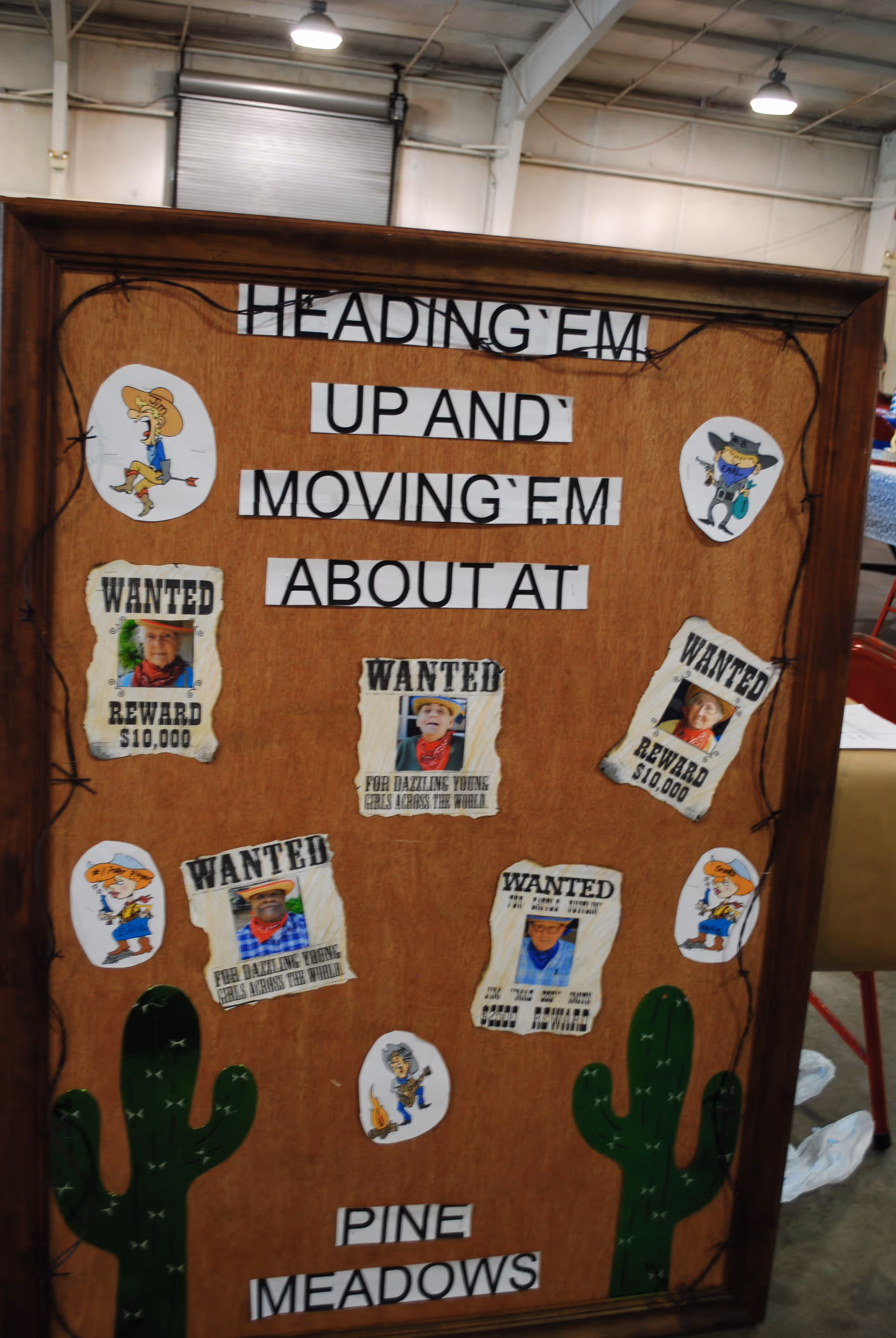 A wooden bulletin board with a western theme featuring the phrase 'HEADING 'EM UP AND MOVING 'EM ABOUT AT PINE MEADOWS'. The board is decorated with cartoon cowboy and cactus cutouts, and several 'WANTED' posters with photos of elderly individuals, some offering a $10,000 reward.
