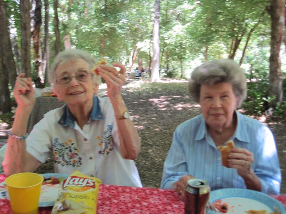 Two elderly women sitting at a picnic table outdoors in a wooded area, enjoying a meal. One woman is holding a sandwich and smiling, while the other is also holding a sandwich. There is a bag of Lay's Classic potato chips, a yellow cup, and a can of soda on the table covered with a red tablecloth.