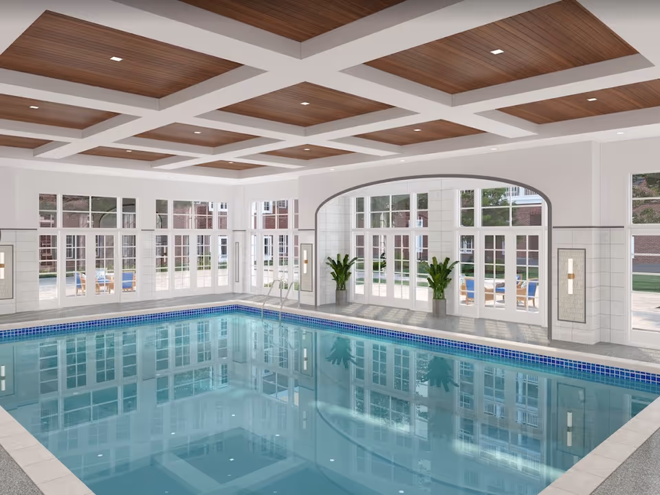 Indoor swimming pool area with large windows and glass doors allowing natural light to enter. The ceiling features wooden panels with recessed lighting. There are two potted plants near the arched glass doors leading outside to a patio with seating. The pool has a tiled edge and a metal ladder for entry.