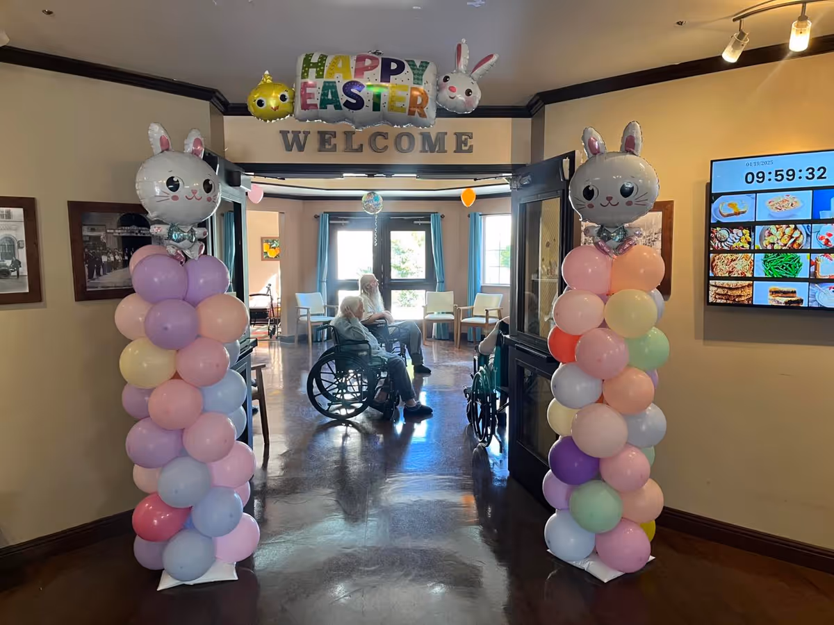 Interior view of a senior living facility decorated for Easter with colorful balloon columns topped with bunny faces on either side of a doorway. Above the doorway is a 'Happy Easter' balloon banner and a 'Welcome' sign. Two elderly individuals in wheelchairs are seated in a well-lit room with chairs and windows in the background. A digital screen displaying meal options and the time is mounted on the right wall.