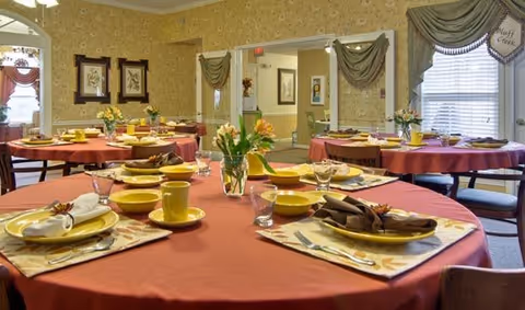 A dining room with round tables covered in red tablecloths, each set with yellow plates, cups, and brown napkins. There are floral centerpieces on the tables, and windows with green drapes. The room has patterned wallpaper and framed artwork on the walls.