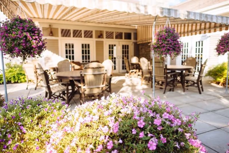 Outdoor patio area with several round tables and wooden chairs under a striped awning. Hanging baskets with purple flowers and flower beds with pink and purple blooms surround the seating area. The background shows a building with multiple glass doors and windows.