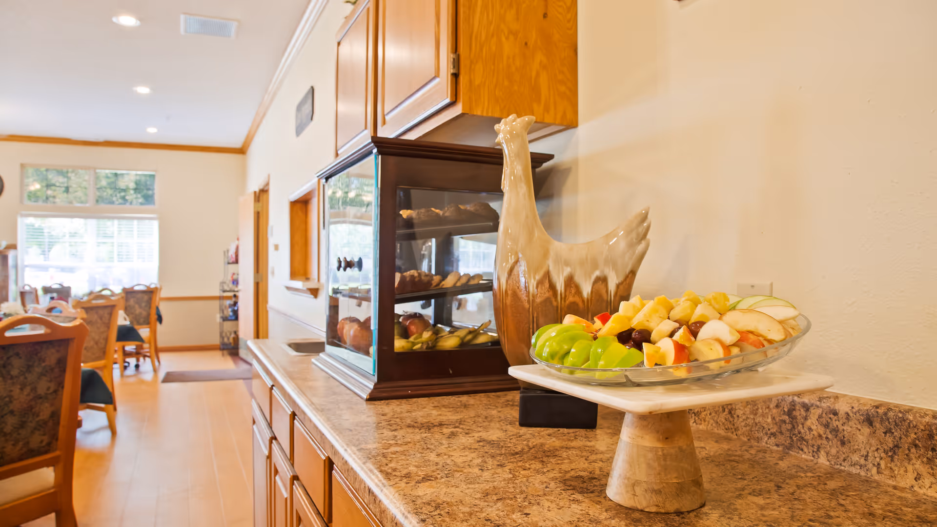 A dining area in a senior living facility with a countertop displaying a decorative ceramic chicken and a platter of assorted fresh fruit. In the background, there are tables and chairs near large windows letting in natural light.