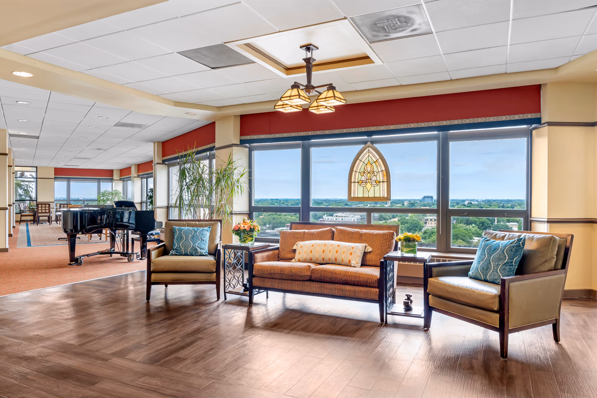 A bright and spacious living room area with large windows offering a panoramic view of greenery and buildings outside. The room features a brown sofa with a decorative pillow, two armchairs with blue patterned cushions, side tables with flower arrangements, and a black grand piano in the background. The ceiling has recessed lighting and a decorative hanging light fixture.