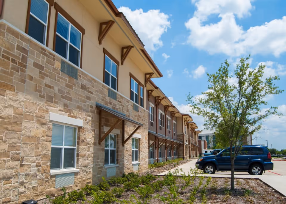 Exterior view of a two-story assisted living facility building with stone and beige siding, multiple windows, a small tree, and parked cars under a partly cloudy blue sky.