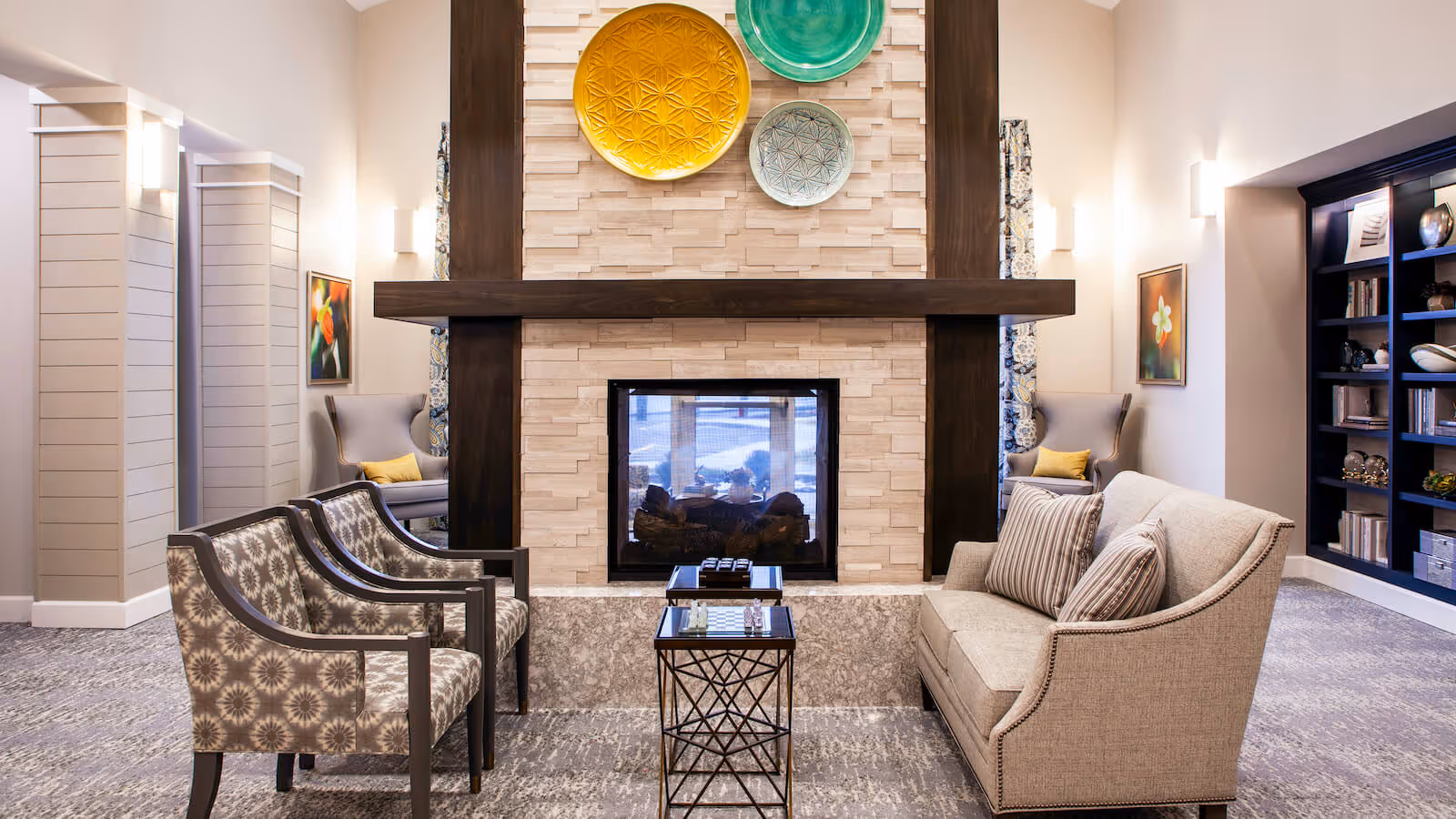 A cozy living room area with a modern fireplace centered on a beige stone wall with dark wooden beams. Above the fireplace are three decorative plates in yellow, green, and light blue. In front of the fireplace are two patterned armchairs on the left and a beige sofa with striped pillows on the right. A small table with a chessboard is placed between the seating. The room has soft lighting, framed floral artwork on the walls, and a dark bookshelf filled with books and decorative items on the right side.