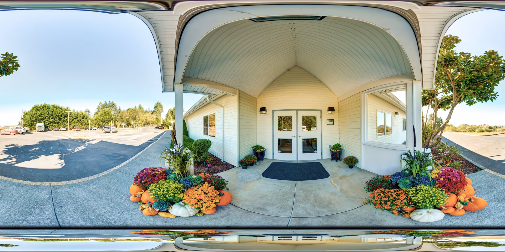 Entrance to Monarch Gardens Memory Care facility with double glass doors under a covered porch. The entrance is decorated with colorful flowers and pumpkins on both sides. There is a parking lot and trees visible in the background under a clear blue sky.