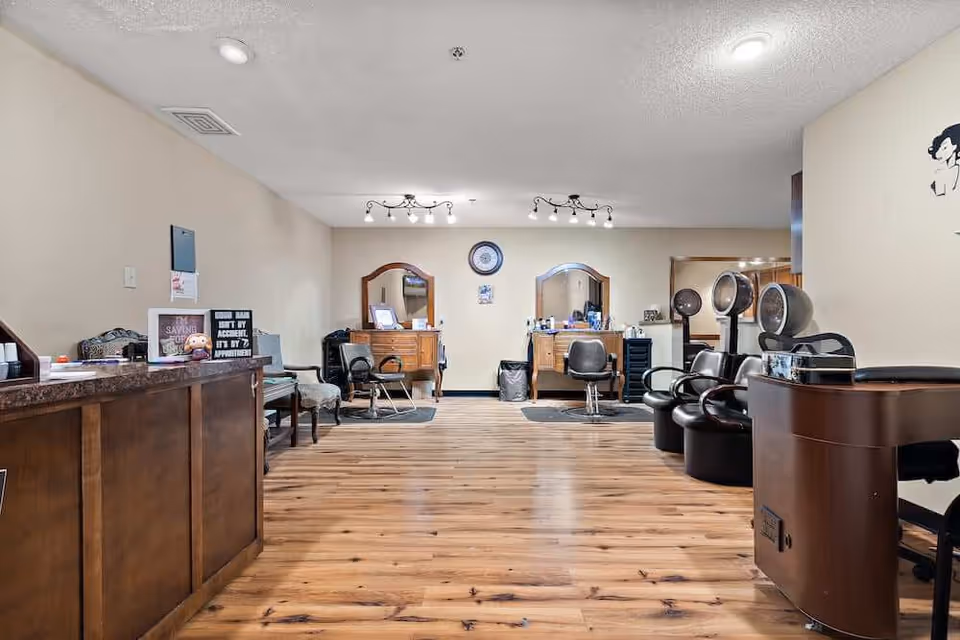 Interior view of a salon area with wooden flooring, two styling stations with mirrors and chairs, hair drying chairs with hooded dryers, and a reception counter on the left side. The walls are beige, and there are ceiling lights illuminating the space.