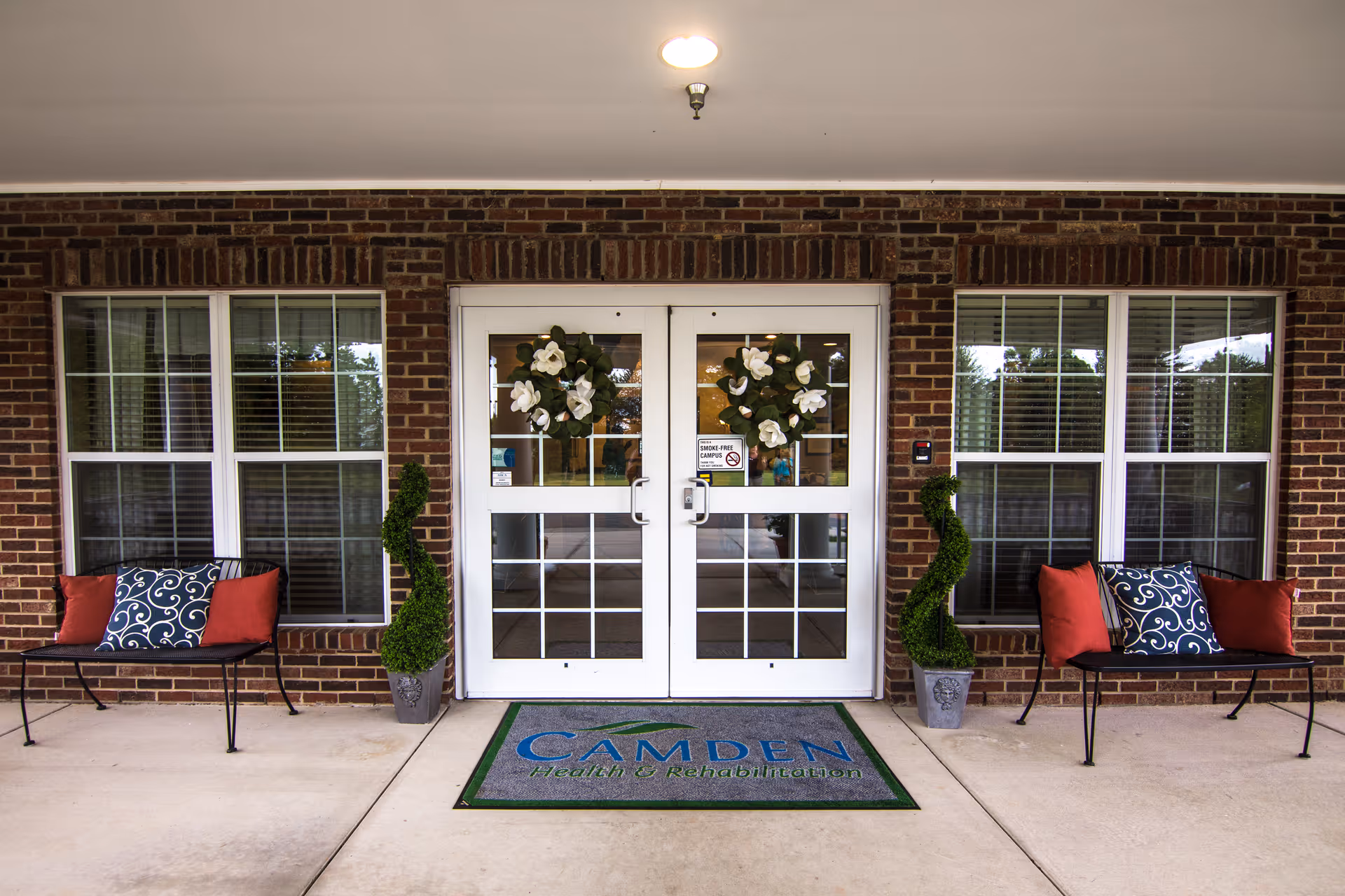 Covered front entrance of Camden Health & Rehabilitation featuring double glass doors with wreaths, flanked by benches, topiary plants and a welcome mat.