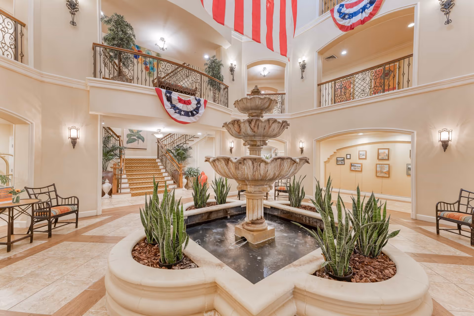 Spacious two-story interior lobby with a central three-tier fountain, surrounding plants, seating, staircases and balcony railings draped with flags.