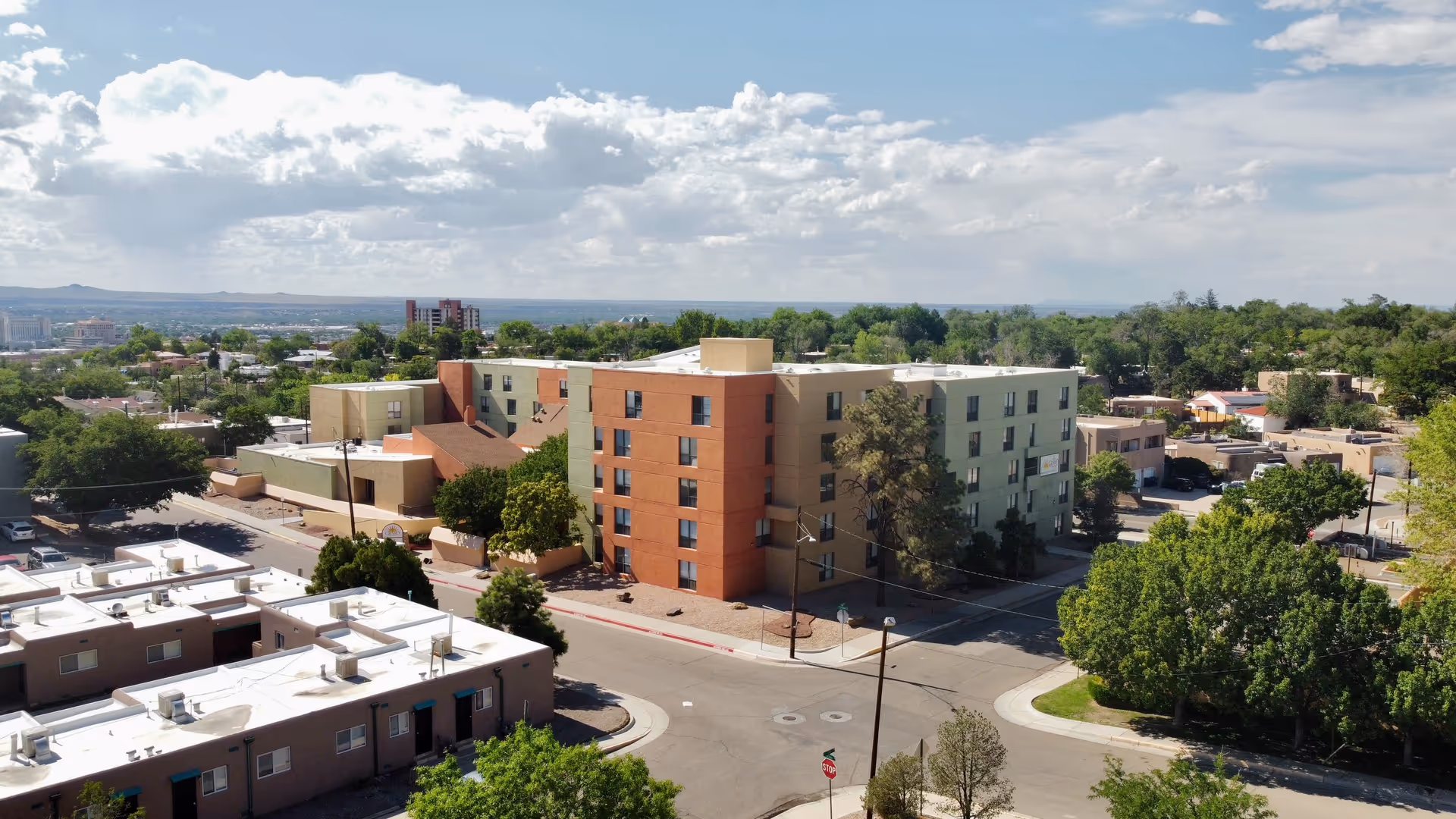 Aerial view of a multi-story senior living facility building surrounded by trees and residential houses under a partly cloudy sky.