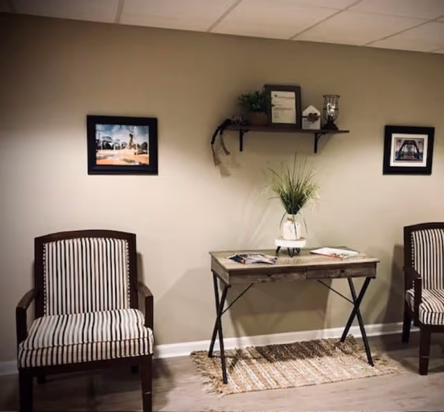 Small sitting area with two striped armchairs, a wooden console table holding a vase of grass and magazines, and framed pictures and a shelf on the wall.