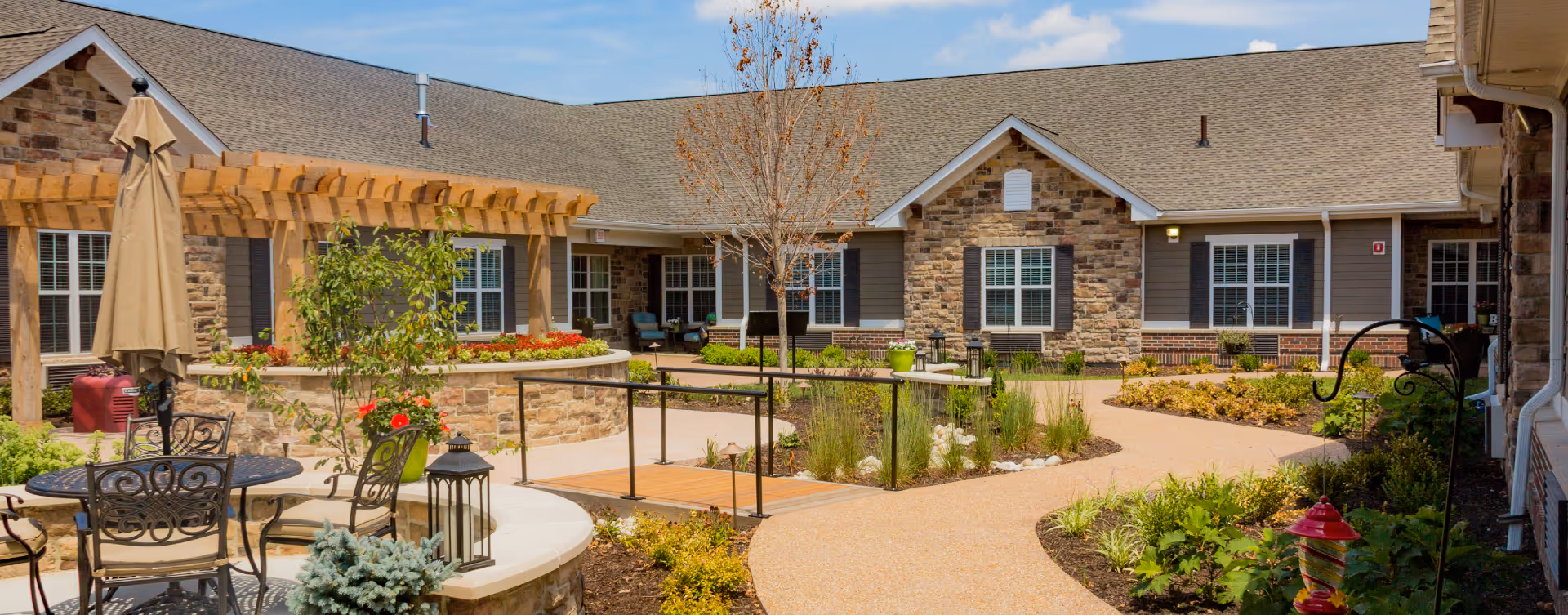 Outdoor courtyard area of a senior living facility with a stone building in the background, a wooden pergola, patio furniture including a table with chairs and an umbrella, landscaped garden beds, a small wooden bridge over a garden area, and a clear blue sky.