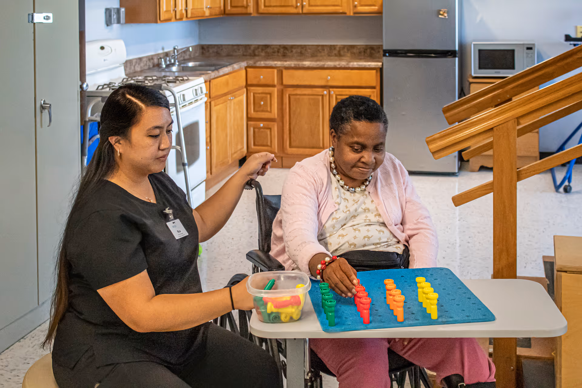 A caregiver assists an elderly woman in a wheelchair as they play a colorful peg game together at a table in a kitchen area with wooden cabinets, a stove, refrigerator, and microwave in the background.