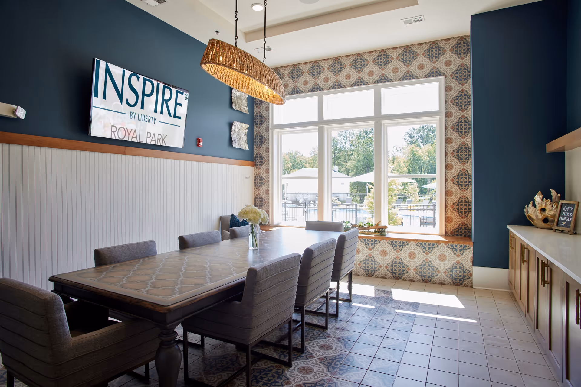 A bright dining room with a long rectangular table surrounded by eight upholstered chairs. The room features a large window with a view of an outdoor area, patterned tile flooring, and a wicker pendant light hanging above the table. The walls are decorated with a combination of dark blue paint and patterned wallpaper. A sign on the wall reads 'INSPIRE BY LIBERTY ROYAL PARK.'