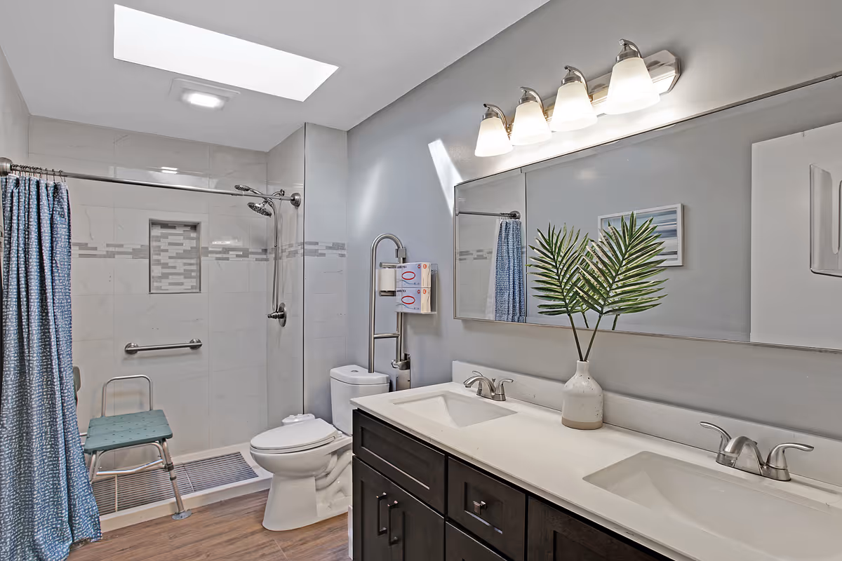 A modern bathroom with a double sink vanity, a large mirror, and a vase with green leaves on the counter. The bathroom features a walk-in shower with a blue shower curtain, a shower chair, grab bars, and a skylight above. The floor has wood-like tiles, and the walls are light gray with white tiles in the shower area.