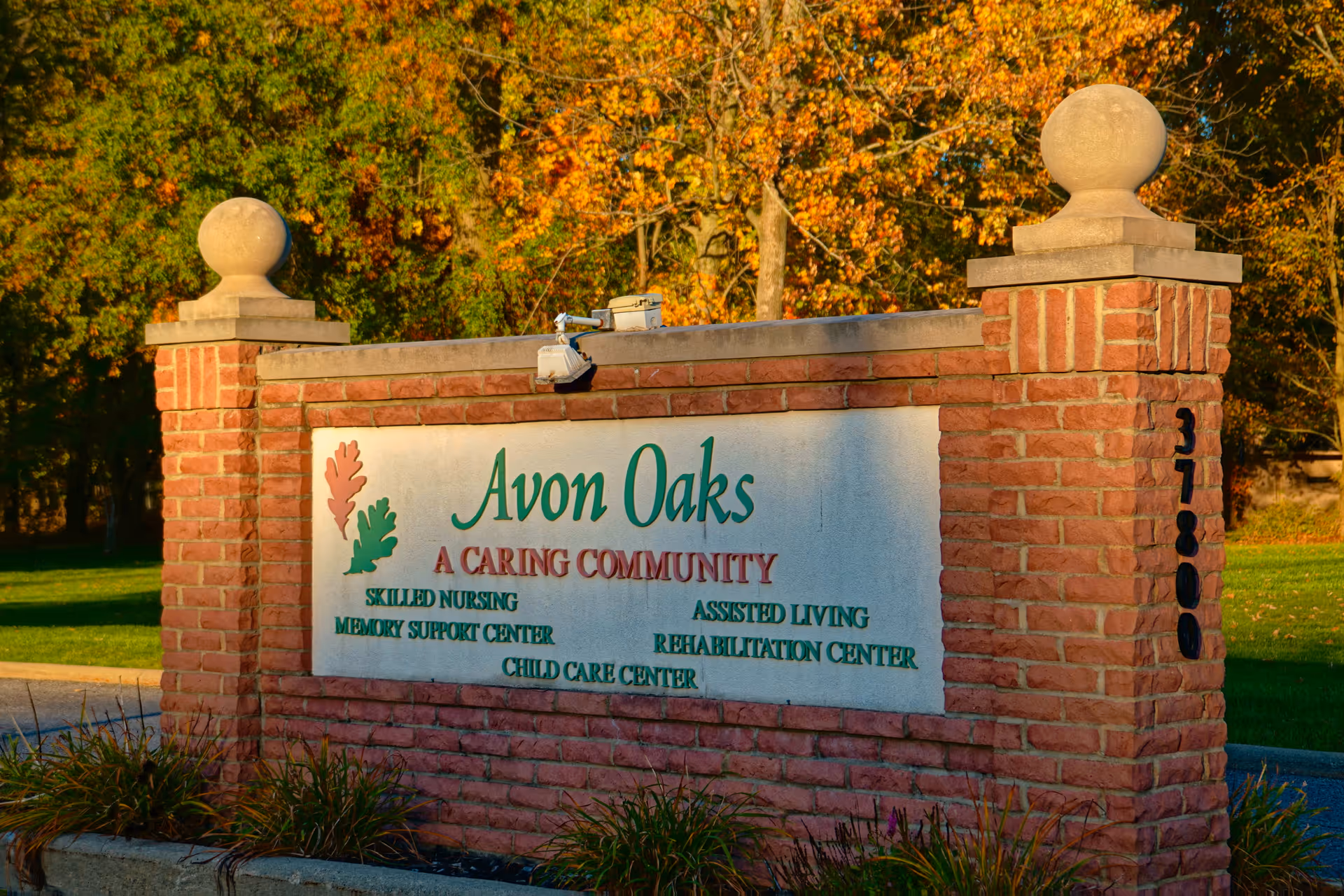 A brick entrance sign for Avon Oaks Caring Community with autumn trees in the background. The sign lists services including skilled nursing, assisted living, memory support center, rehabilitation center, and child care center.