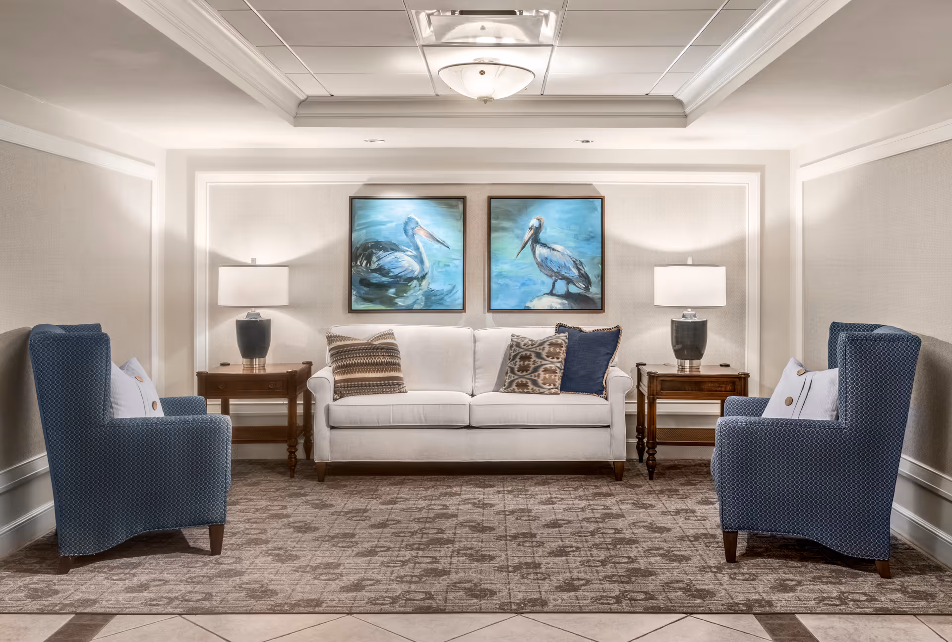 A well-lit living room area with a white sofa adorned with patterned and solid pillows, flanked by two wooden side tables each holding a lamp. Two blue upholstered armchairs face the sofa. Above the sofa, there are two framed paintings of pelicans on a blue background. The room features a patterned carpet and a tray ceiling with a central light fixture.