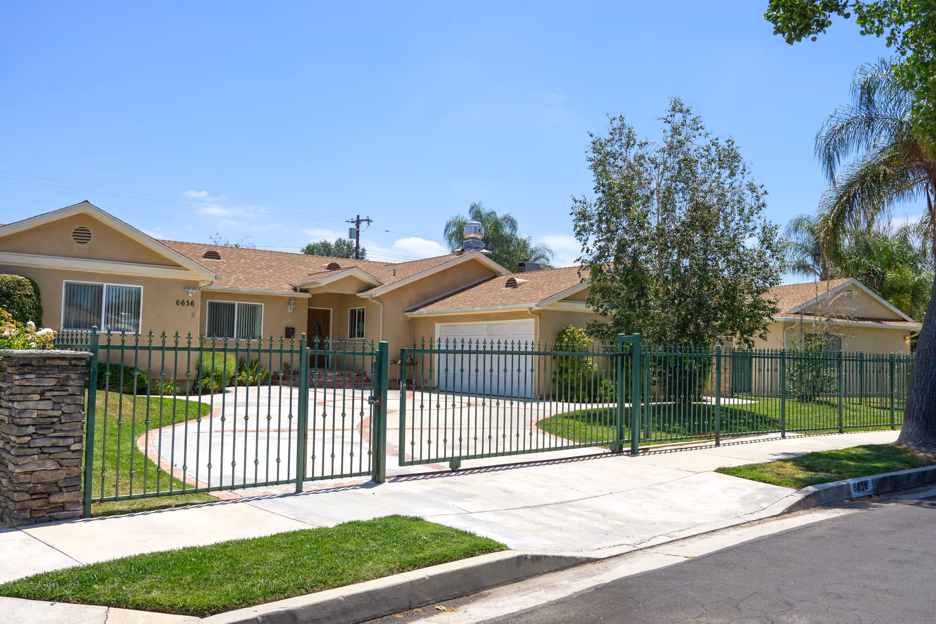 Exterior view of a single-story residential building with a beige facade and a brown shingle roof, surrounded by a green metal fence. There is a driveway leading to a double garage, a tree in the front yard, and a clear blue sky overhead.