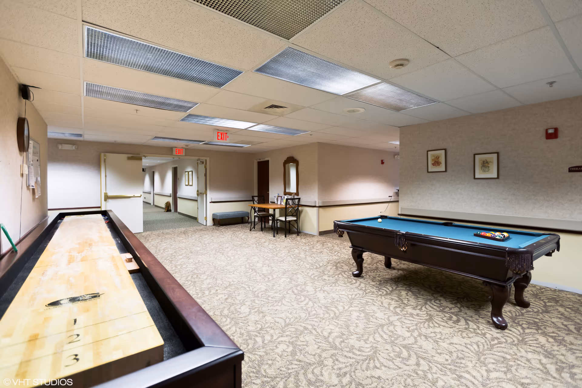 A recreational room in a senior living facility featuring a shuffleboard table on the left and a pool table on the right. There is a small table with four chairs and a mirror on the far wall. The room has patterned carpet, beige walls, and a drop ceiling with fluorescent lighting. An open doorway leads to a hallway with exit signs visible.