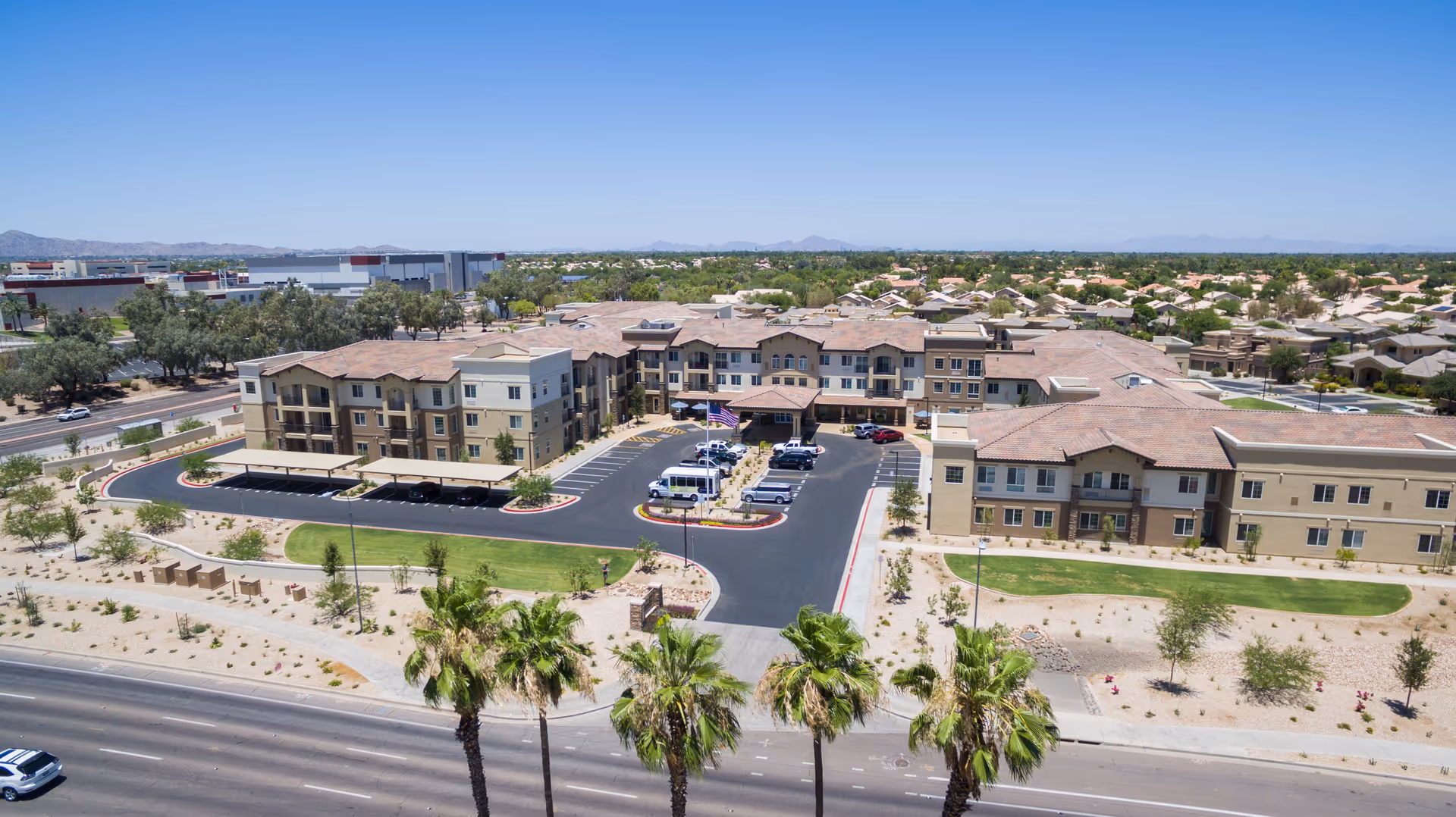Aerial view of The Enclave at Chandler Senior Living facility showing a large, multi-story building with beige and tan exterior, surrounded by landscaped grounds with palm trees and desert plants. There is a parking lot with several vehicles and a driveway leading to the main entrance. The surrounding area includes residential neighborhoods and distant mountains under a clear blue sky.