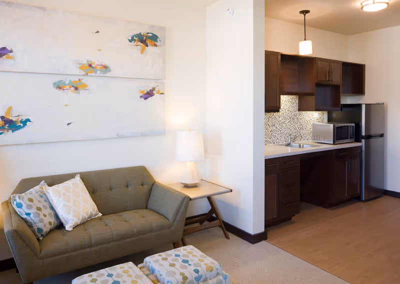 A compact living area with a green sofa, patterned pillows and ottomans, a side table with lamp, wall art, and a small kitchenette with dark cabinets and stainless appliances.