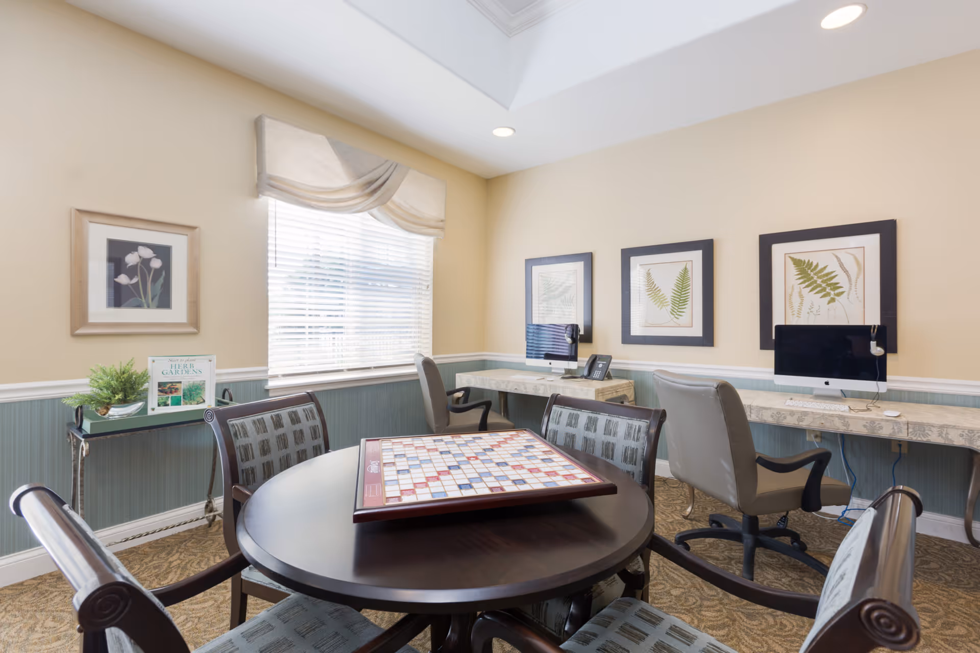 Bright communal activity room featuring a round table with a Scrabble board, surrounding chairs, and a wall of desks with computers and framed botanical artwork.