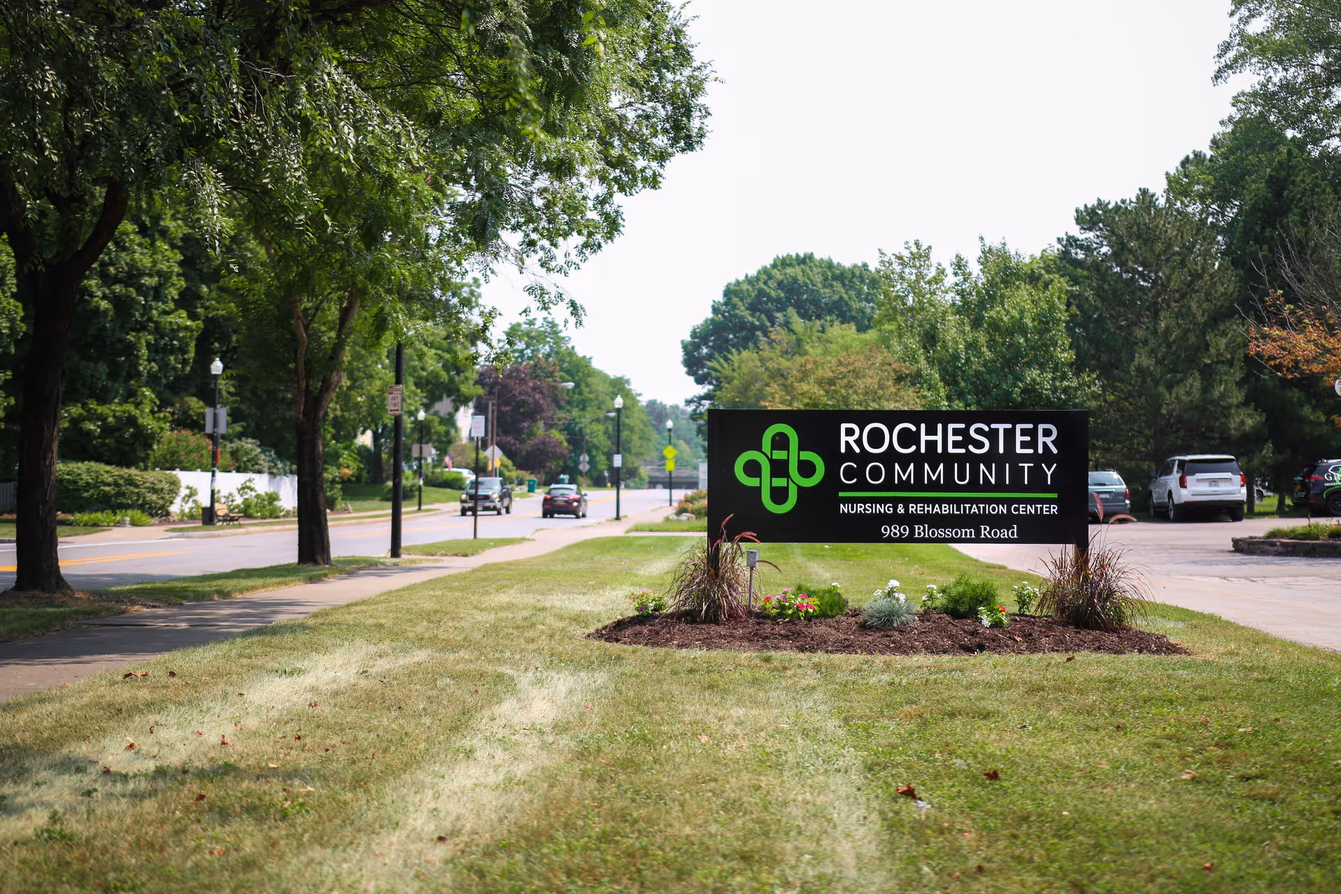 A lawn and roadside sign reading "Rochester Community Nursing & Rehabilitation Center 989 Blossom Road" with trees and parked cars in the background.