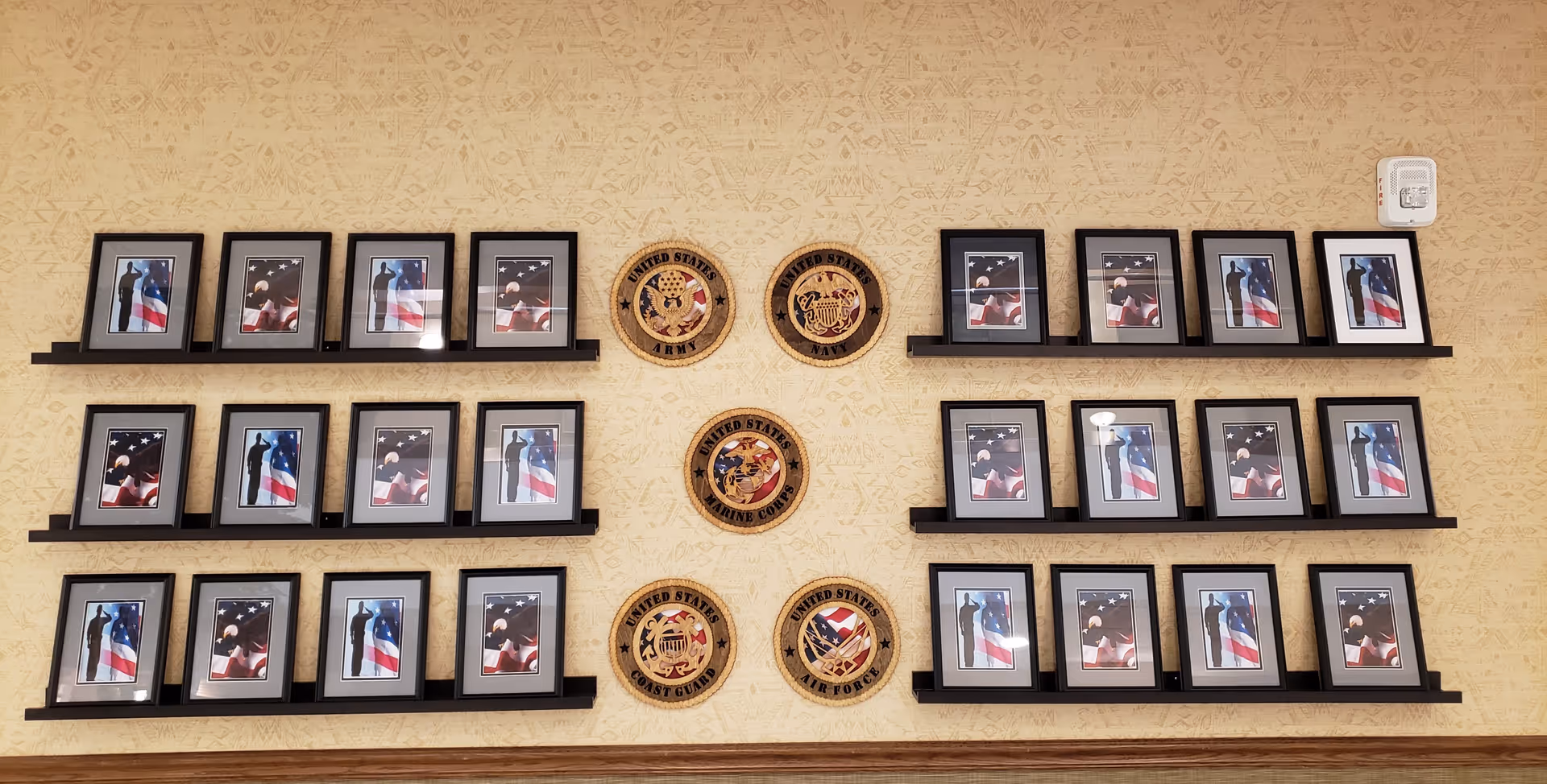 A wall display featuring multiple framed photographs of a silhouette saluting in front of an American flag, arranged on black shelves. In the center of the display are five circular wooden plaques representing the United States Army, Navy, Marine Corps, Coast Guard, and Air Force. The wall has a beige patterned wallpaper background.
