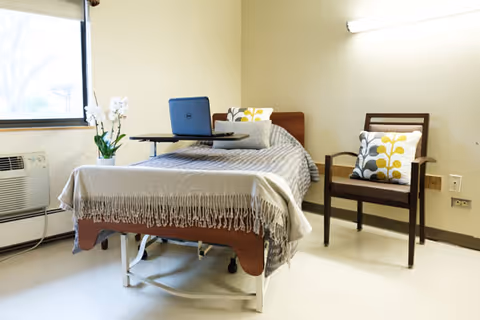 A neatly made hospital-style bed with a gray blanket and decorative pillows in a simple, clean room. A small table with a laptop is placed on the bed. Next to the bed is a wooden chair with a cushion featuring a yellow and gray leaf pattern. A potted white orchid is on the windowsill beside an air conditioning unit. The walls are light beige and the floor is light-colored.