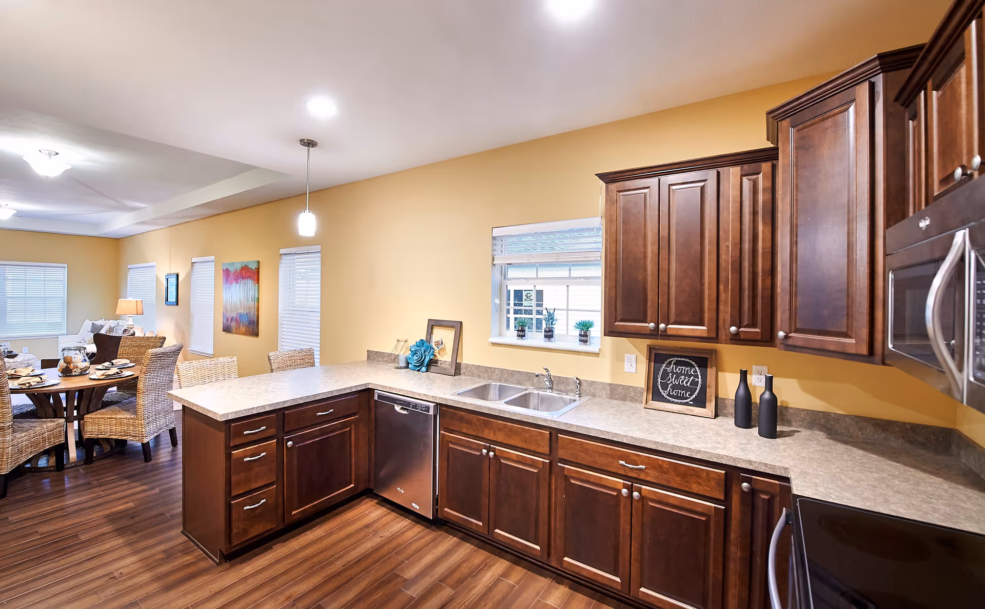 A modern kitchen with dark wood cabinets, a double sink under a window, a dishwasher, and a microwave. The kitchen opens into a dining area with a round table and wicker chairs. The walls are painted yellow, and the floor is a dark wood laminate.