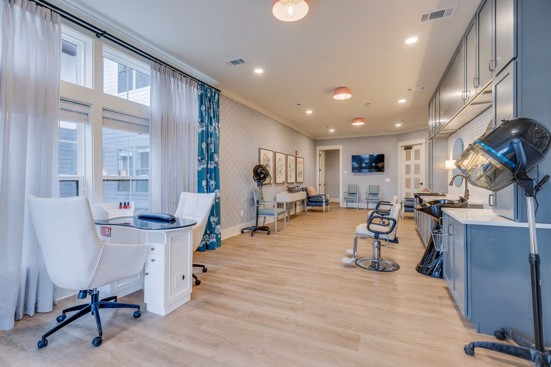 A bright and spacious salon area with large windows covered by sheer white and blue floral curtains. The room features white swivel chairs at manicure tables on the left, and black salon chairs with hair dryers on the right. The floor is light wood, and the walls are decorated with framed artwork and patterned wallpaper. There is a television mounted on the far wall and additional seating along the back wall.